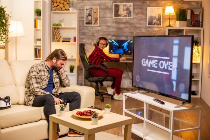 Homem sentado na sala decepcionado com o game over no video game. Mulher sentada ao fundo no computador olhando para ele.